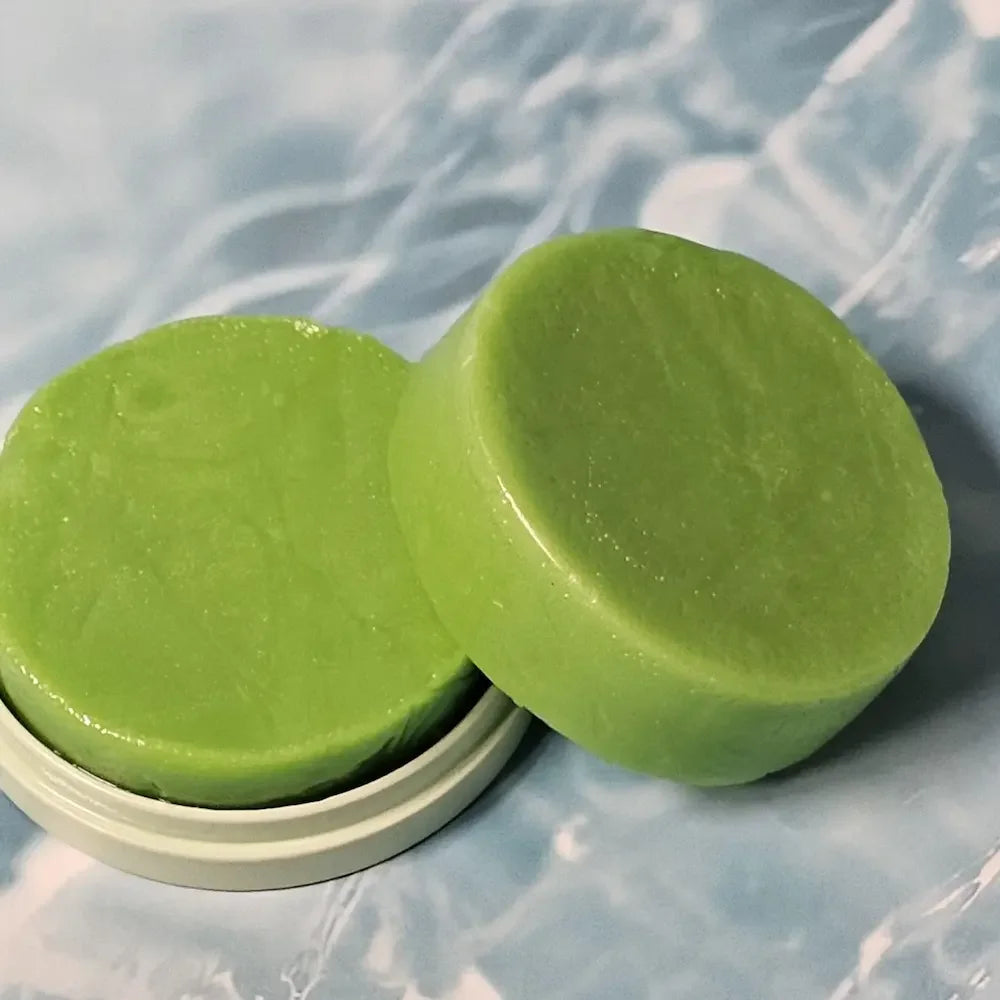 Two round green shampoo bars placed on a tin container with a soft, light blue marbled background.